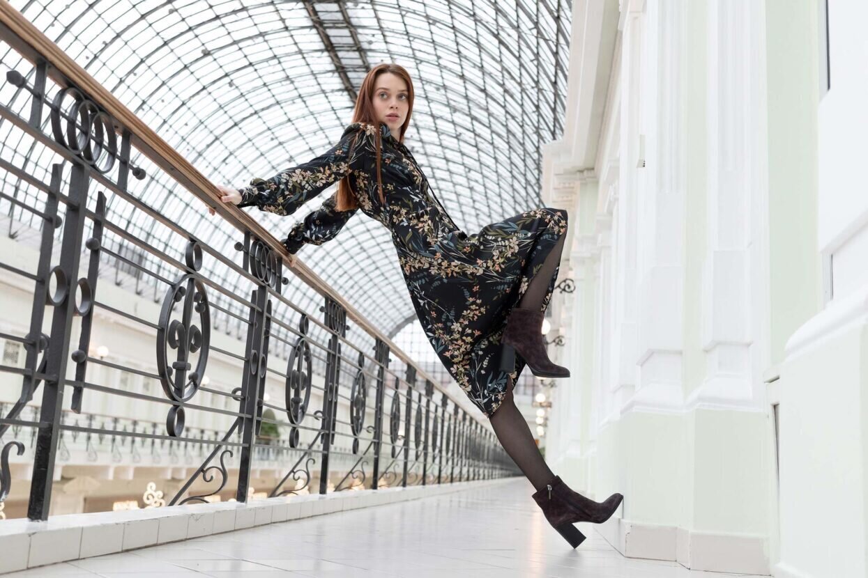 Conceptual posing. European beautiful girl with long red hair in a black dress with a floral print and batilions leans on railing in a strange position